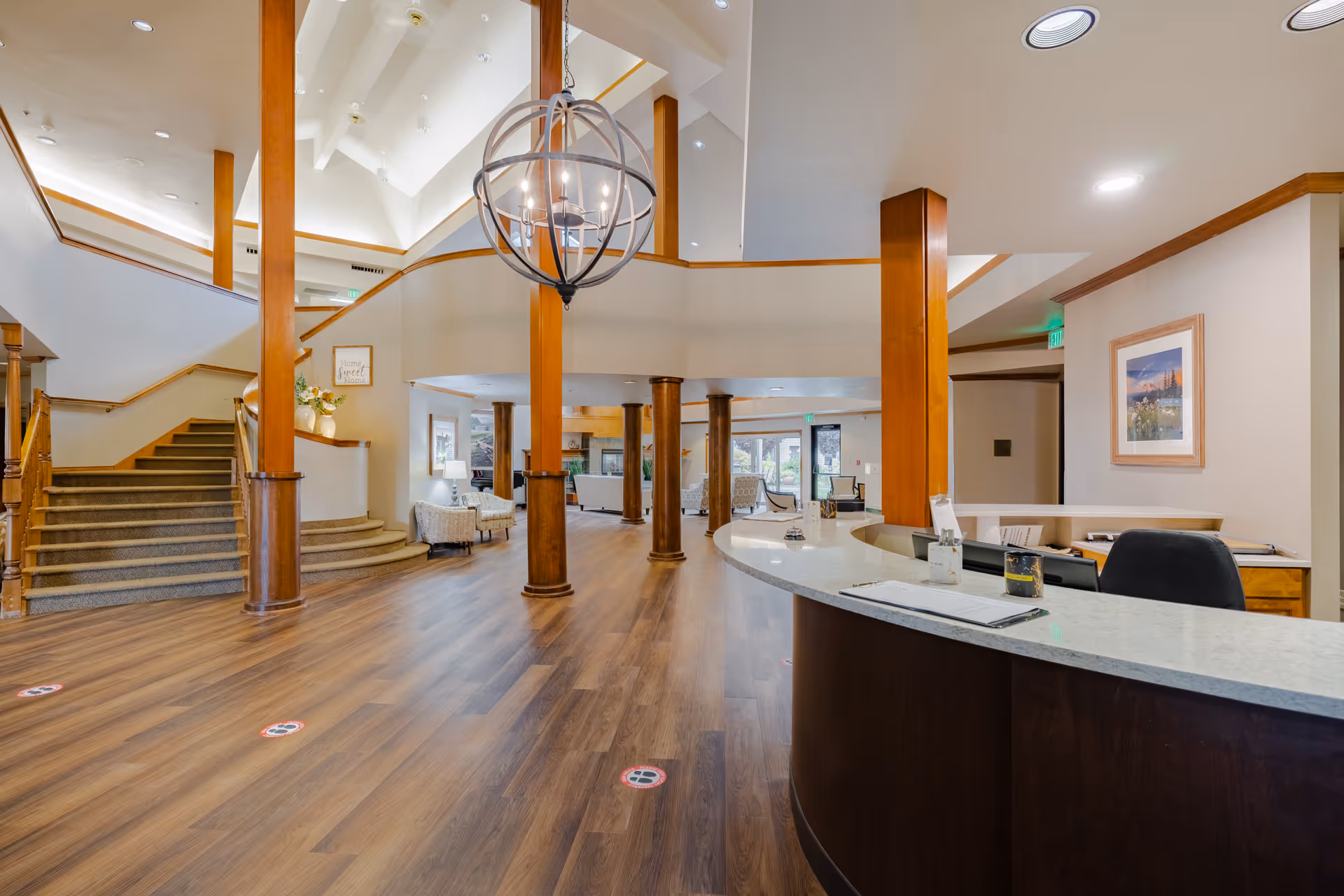 Spacious senior living facility lobby with wooden flooring, a curved reception desk on the right, wooden pillars, a modern spherical chandelier hanging from the ceiling, and a carpeted staircase on the left. Comfortable seating areas with armchairs and lamps are visible in the background near large windows.