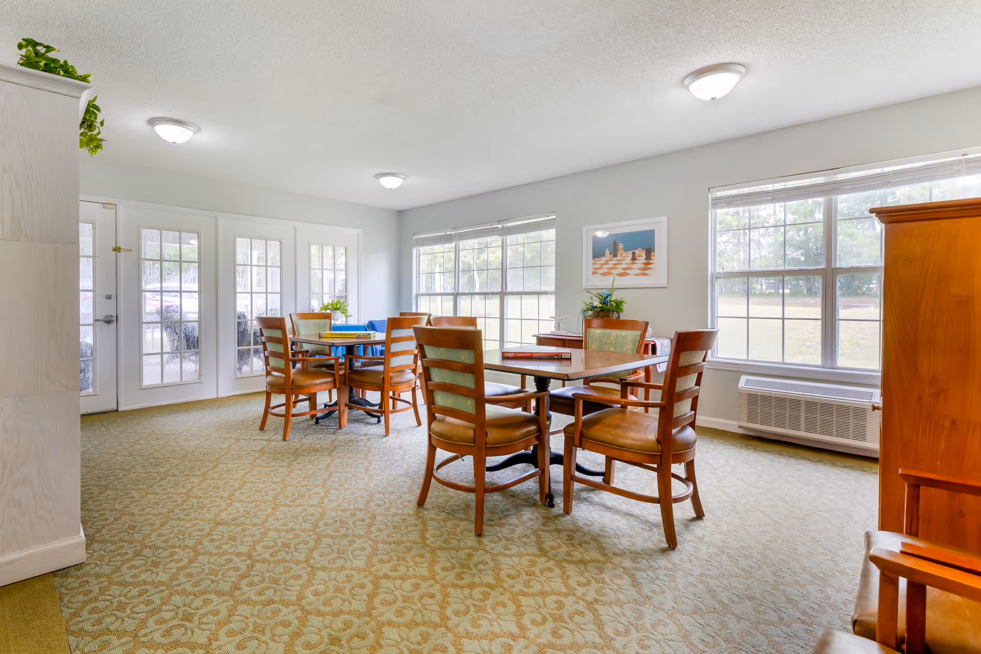 A bright room with large windows letting in natural light. The room has two tables with wooden chairs around them. One table has a board game on it. The walls are light-colored, and there is a framed picture on the wall. The floor is carpeted with a patterned design. There are ceiling lights and a white door with glass panels leading outside.