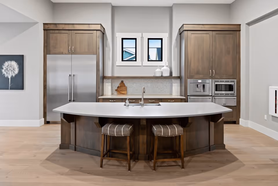 Modern kitchen with a large white island countertop featuring a built-in sink and two cushioned stools. The kitchen has wooden cabinets, a stainless steel refrigerator, built-in microwave and oven, and two small windows above the countertop. Light wood flooring and neutral-colored walls complete the space.
