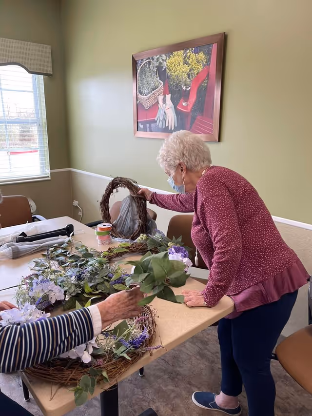 Two elderly women wearing masks are engaged in a craft activity at a table, arranging artificial flowers and greenery on grapevine wreaths in a well-lit room with green walls and a framed picture of gardening gloves and flowers on the wall.