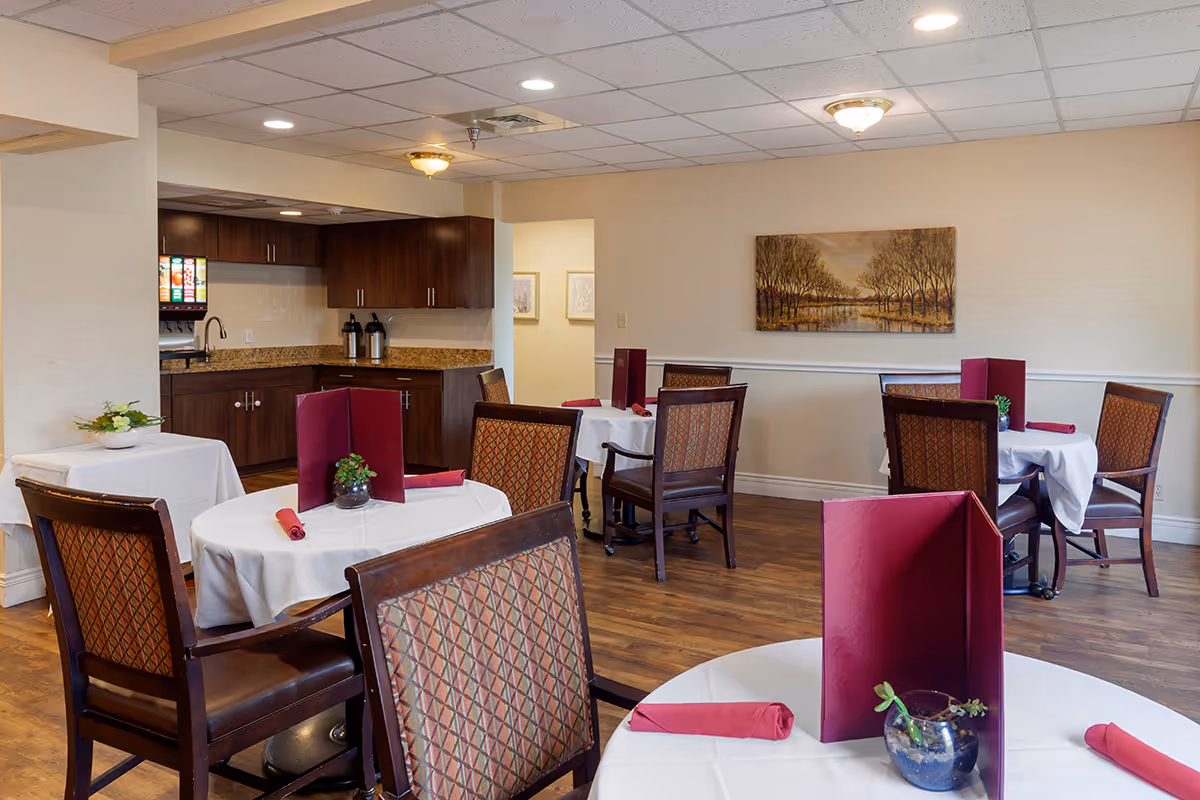 A dining room in an assisted living facility with round tables covered in white tablecloths, each set with red napkins and red menu holders. The room has wooden chairs with patterned upholstery, wood flooring, and a kitchenette area with dark wood cabinets and a soda fountain machine. A landscape painting hangs on the beige wall.