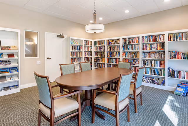 A cozy interior reading room with an oval wooden table surrounded by six chairs and bookshelves lining the walls.