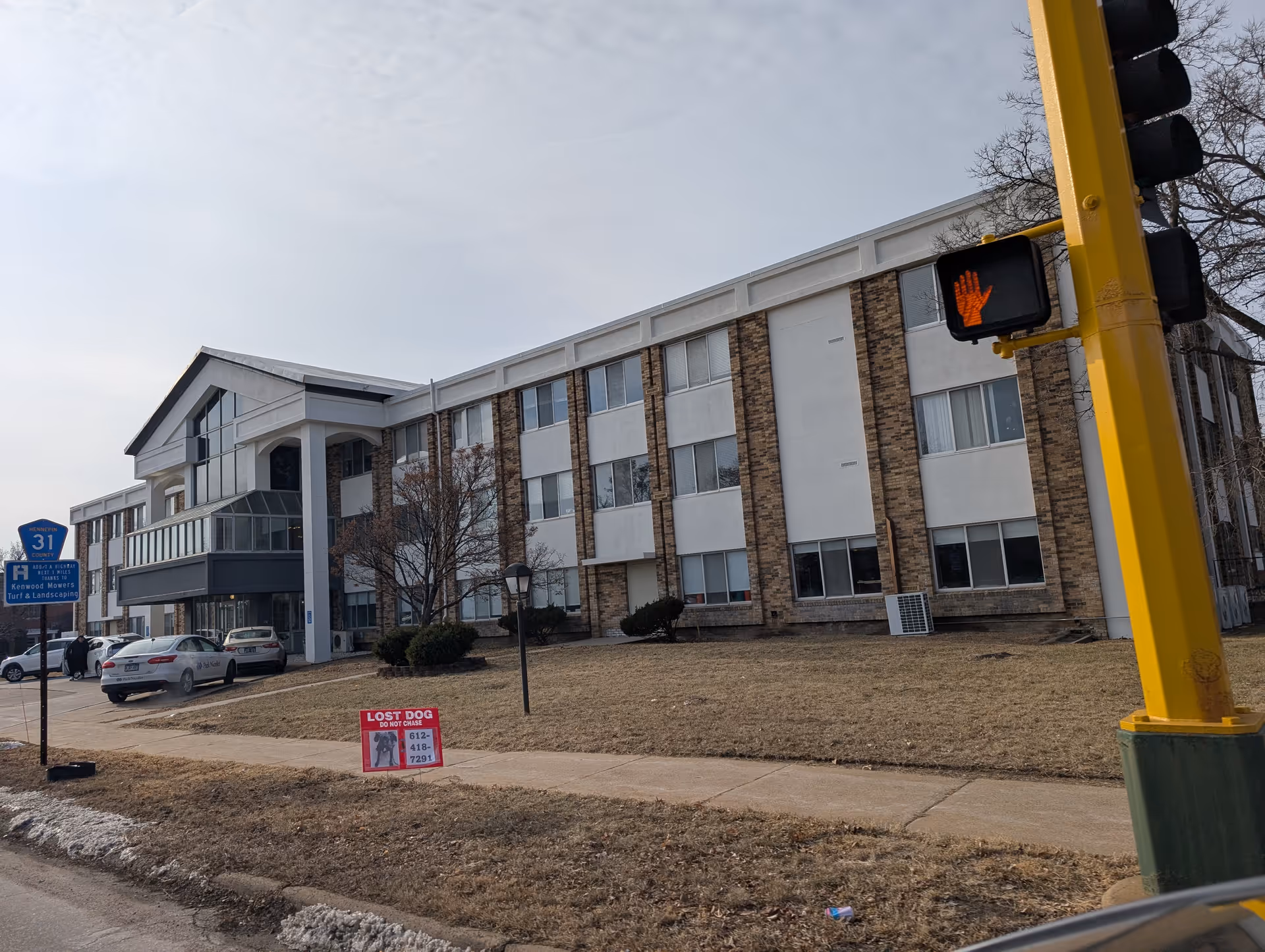 Exterior view of a three-story building with a combination of brick and white paneling. There are several windows, a small parking area with cars, a sidewalk, and a traffic light pole with a pedestrian stop signal. A red lost dog sign is placed on the grass near the sidewalk.