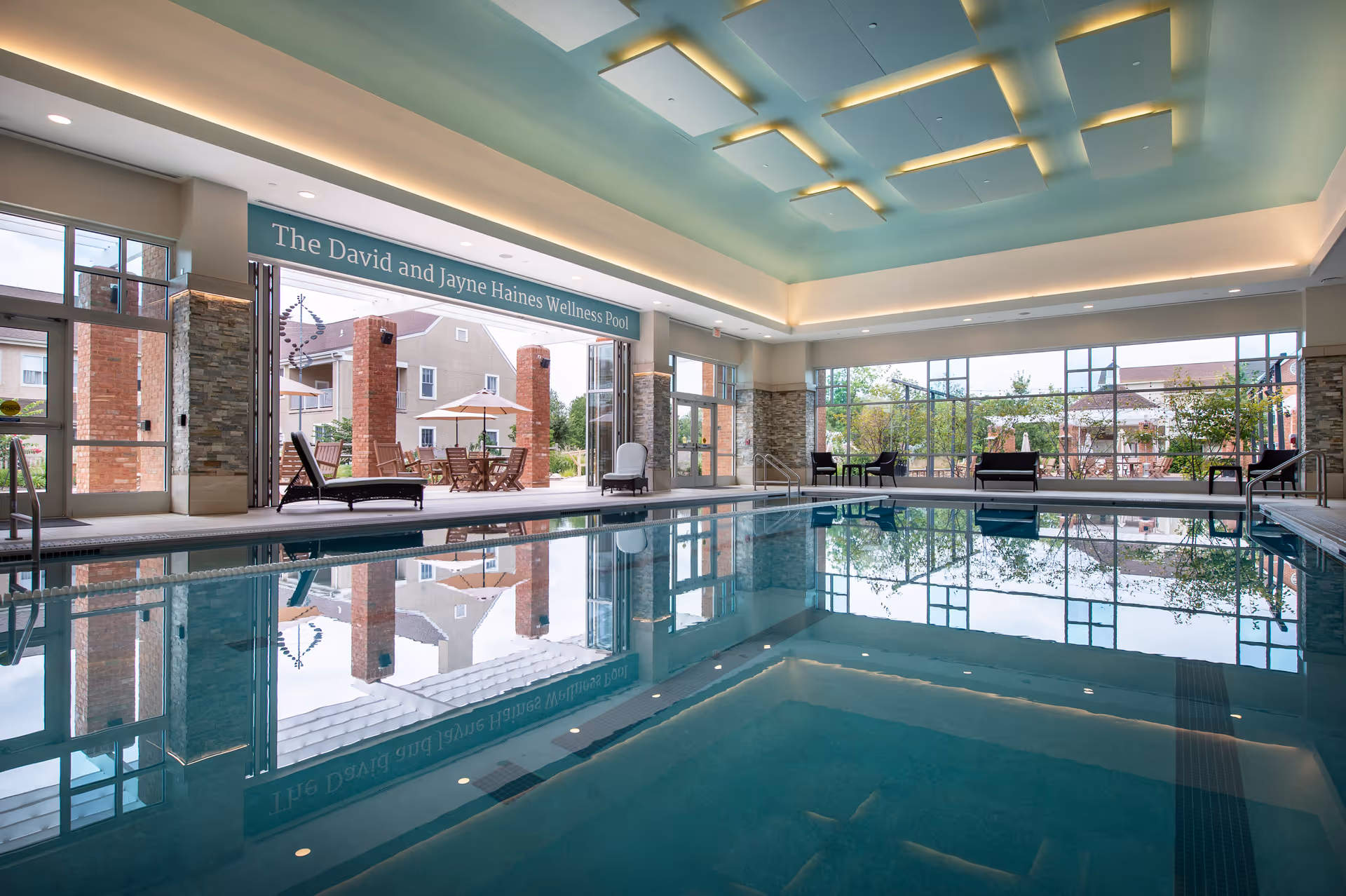Indoor swimming pool area with large windows and glass doors opening to an outdoor patio with tables, chairs, and umbrellas. The ceiling has recessed lighting and a modern design. A sign above the doors reads 'The David and Jayne Haines Wellness Pool.'