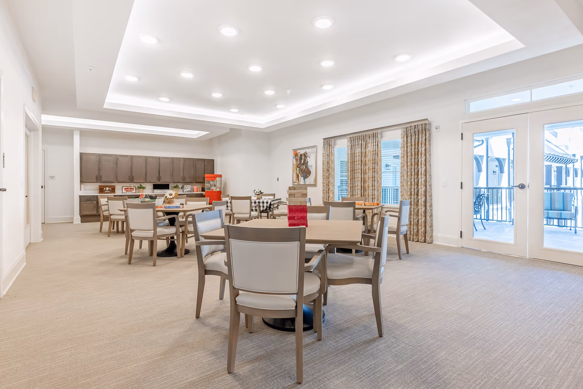 A bright and spacious community room with several tables and chairs arranged for group activities. The room features a popcorn machine, cabinets along the back wall, and large windows with patterned curtains letting in natural light. There is a double glass door leading to an outdoor patio area with seating.