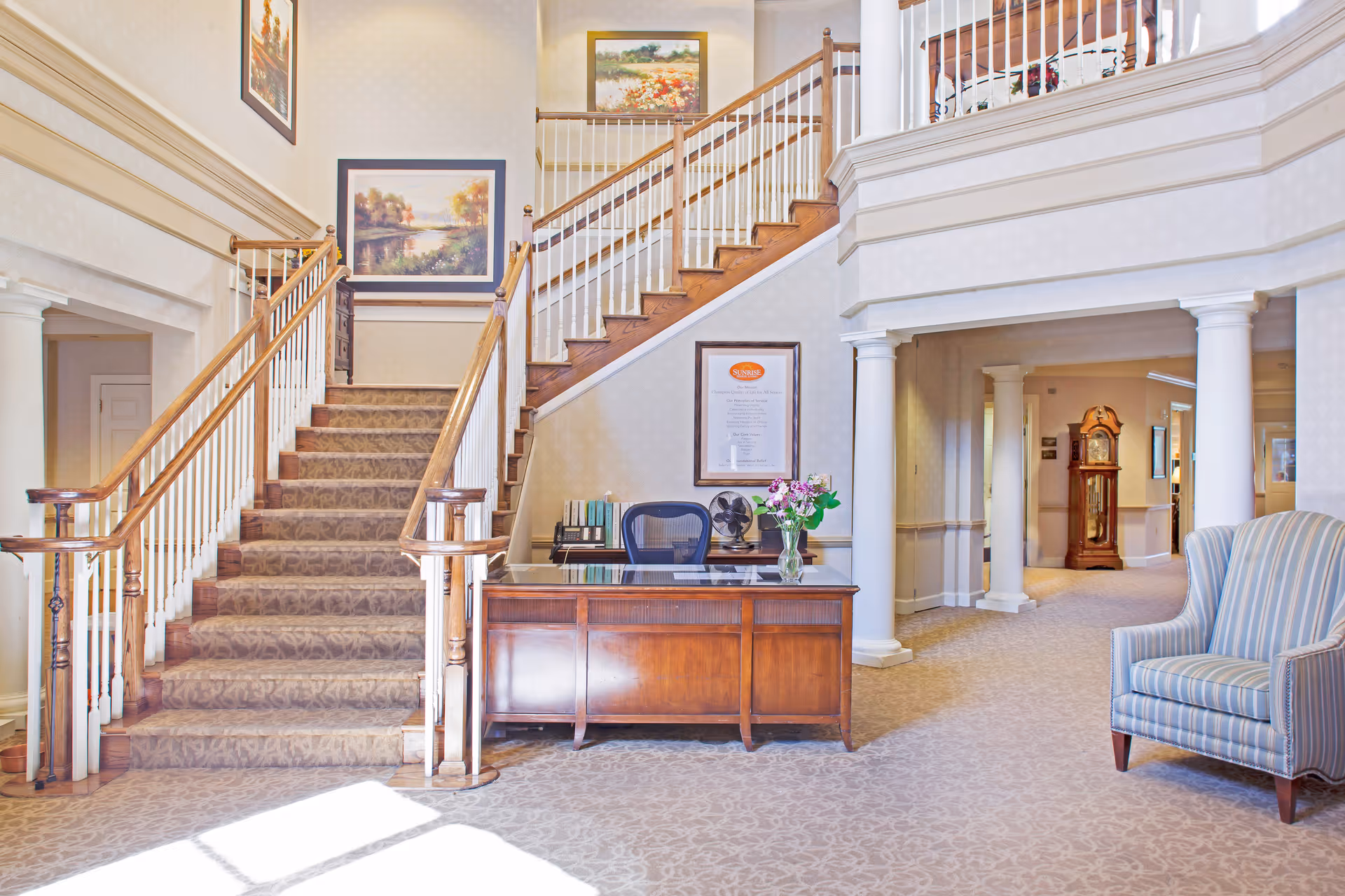 Interior view of a senior living facility lobby with a wooden staircase carpeted in a patterned fabric, a wooden reception desk with a chair and a vase of flowers, framed paintings on the walls, white columns, and a striped armchair on the right side.