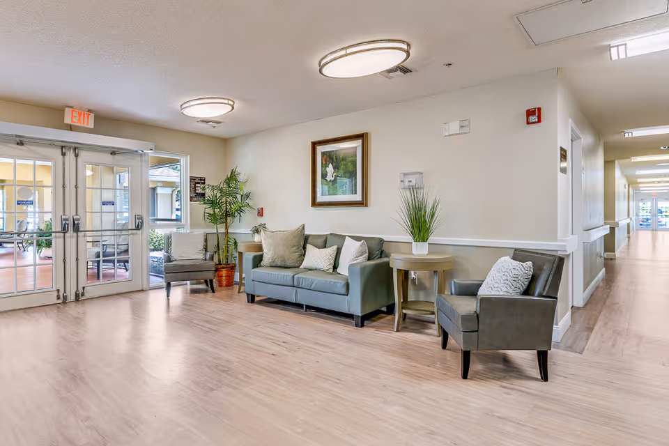 A bright and spacious senior living facility lobby area with light wood flooring, a gray sofa with cushions, two gray armchairs, a round wooden side table with a potted plant, a framed picture on the wall, and glass double doors leading outside. There is also a potted plant near the sofa and ceiling lights illuminating the space.