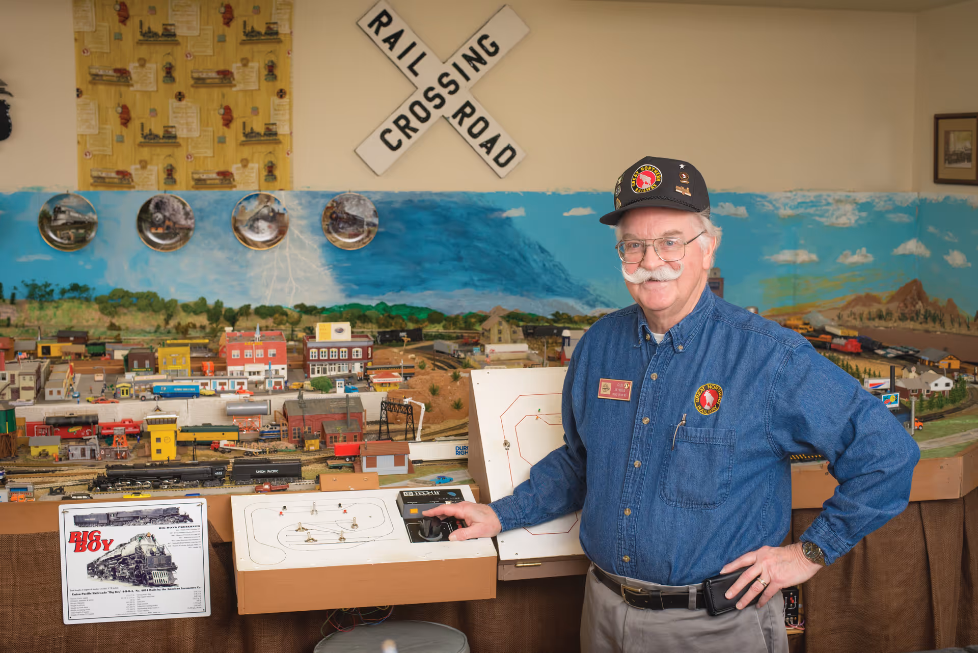 An elderly man with a mustache and glasses wearing a blue shirt and a cap stands next to a detailed model train setup. The background features a painted mural of a landscape with hills and sky, decorative plates on the wall, and a railroad crossing sign. The man is smiling and appears to be controlling the model train with a control panel.