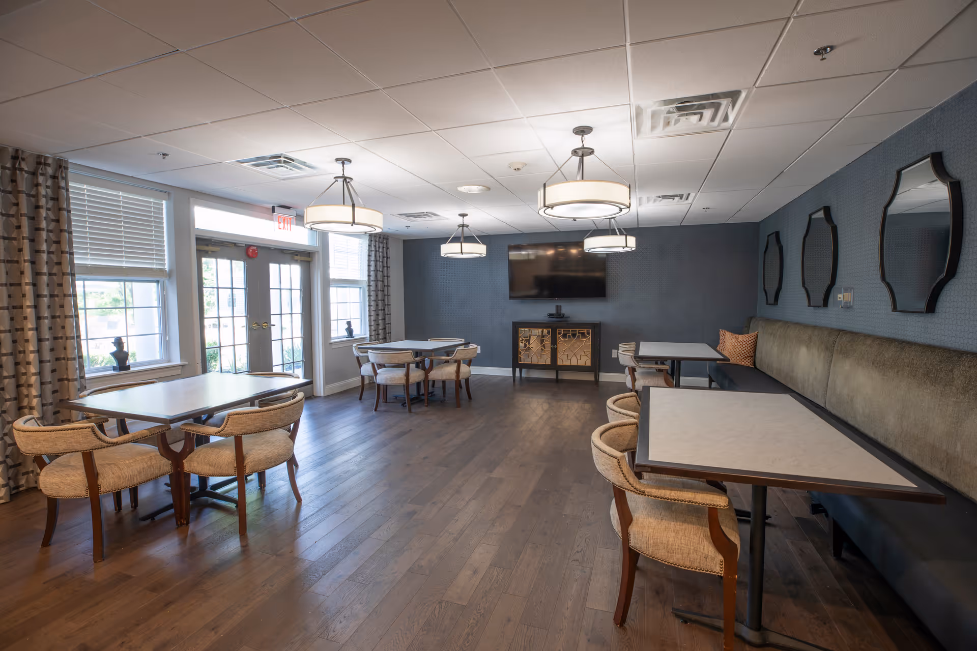A well-lit dining room with wooden floors, several tables with chairs, a long cushioned bench along one wall, decorative mirrors above the bench, large windows with patterned curtains, and a wall-mounted flat-screen TV above a cabinet.