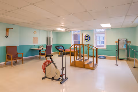 A bright rehabilitation room with exercise equipment including a stationary bike, treadmill, parallel bars, and wooden practice stairs.