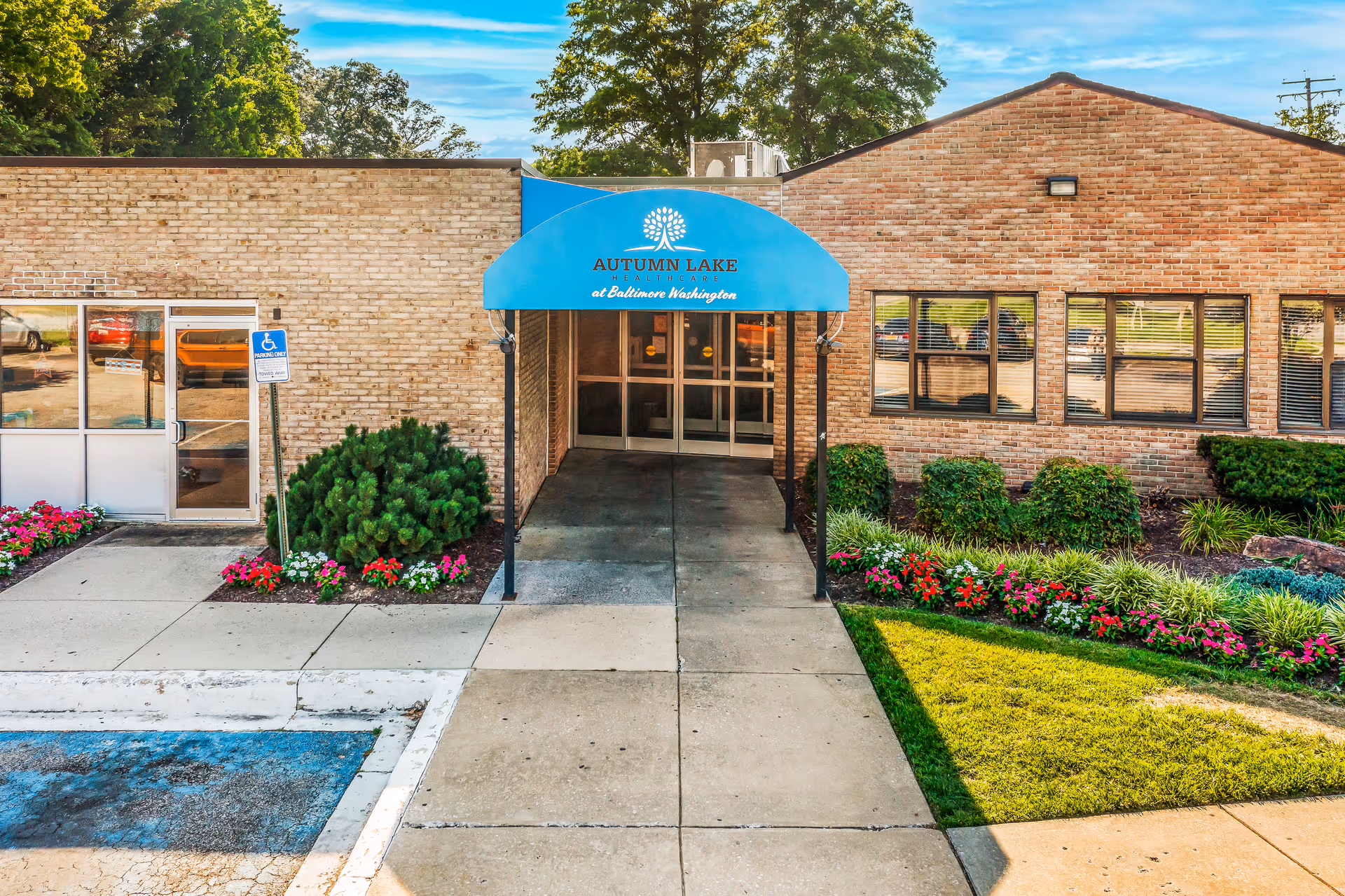 Entrance of Autumn Lake Healthcare at Baltimore Washington with a blue awning displaying the facility's name. The building is made of light brown brick with windows on the right side. There are flower beds with colorful flowers and green shrubs on either side of the walkway leading to the entrance. A handicapped parking space is visible in the foreground on the left.