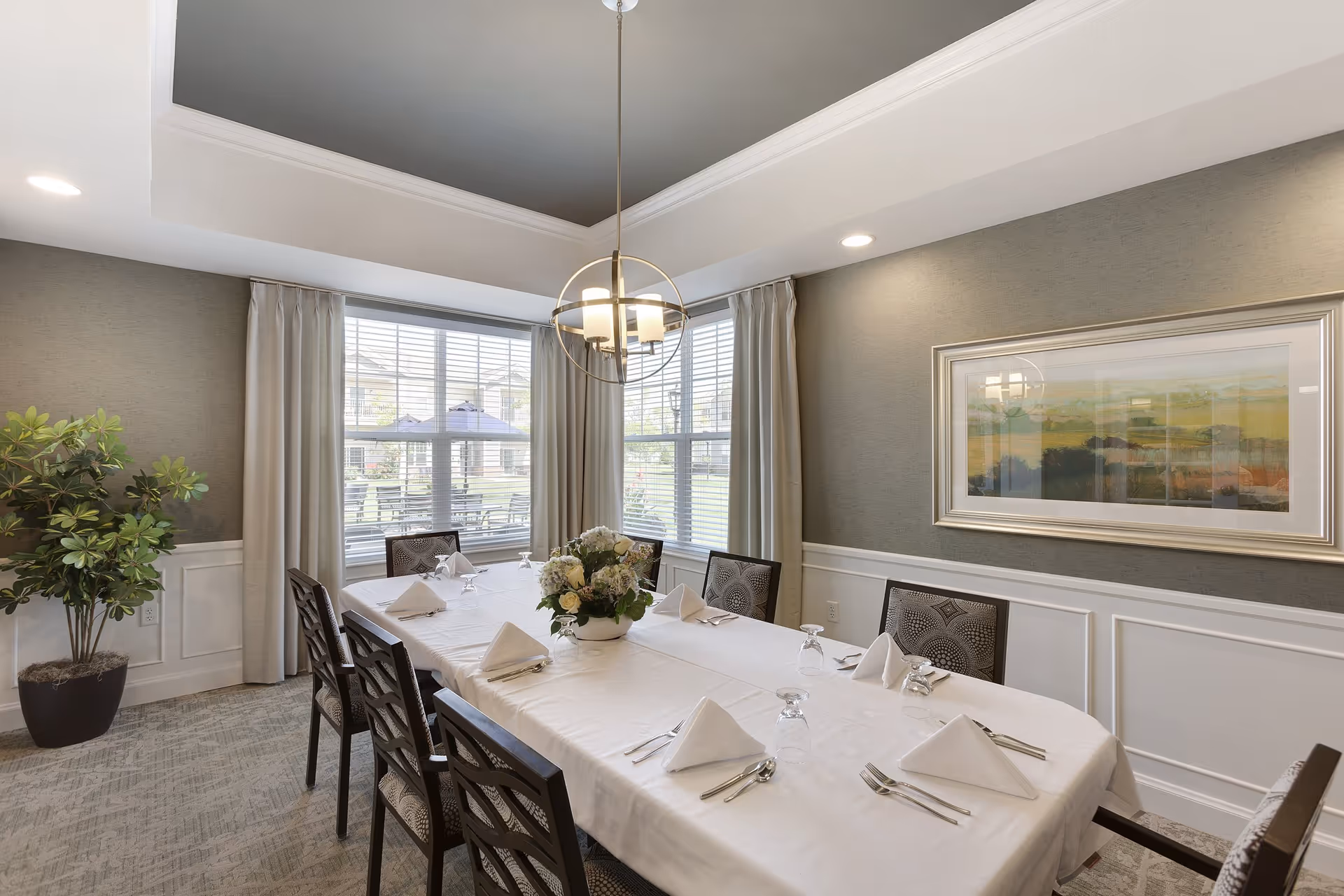 A formal dining room with a rectangular table covered with a white tablecloth, set with folded white napkins, silverware, and upside-down glasses. A floral centerpiece is in the middle of the table. The room has large windows with white blinds and beige curtains, a modern chandelier hanging from a tray ceiling, a potted plant in the corner, and a framed landscape painting on the wall.