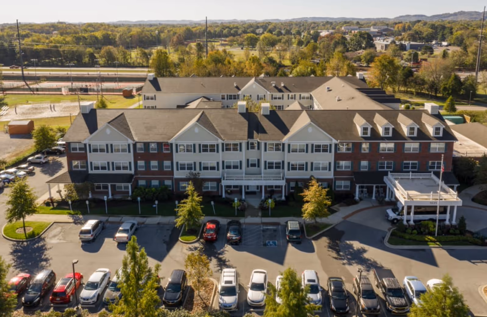 Aerial view of a large senior living facility building named Vitality Living Franklin, surrounded by a parking lot with multiple cars and green trees. The building has a multi-story structure with a combination of white and brick exterior walls, multiple windows, and a covered entrance area with an American flag. The background shows a landscape of trees and distant hills under a clear sky.