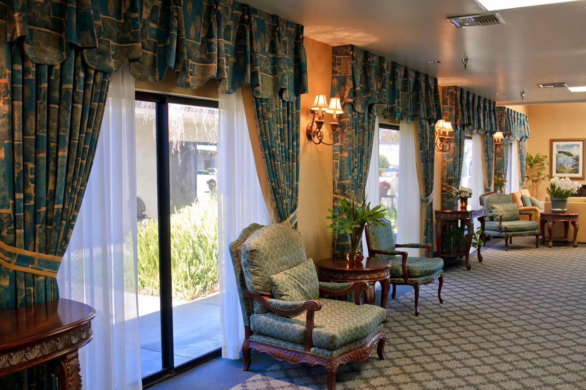 A bright lounge with upholstered armchairs, wooden side tables, wall sconces, and large curtained windows and sliding doors.