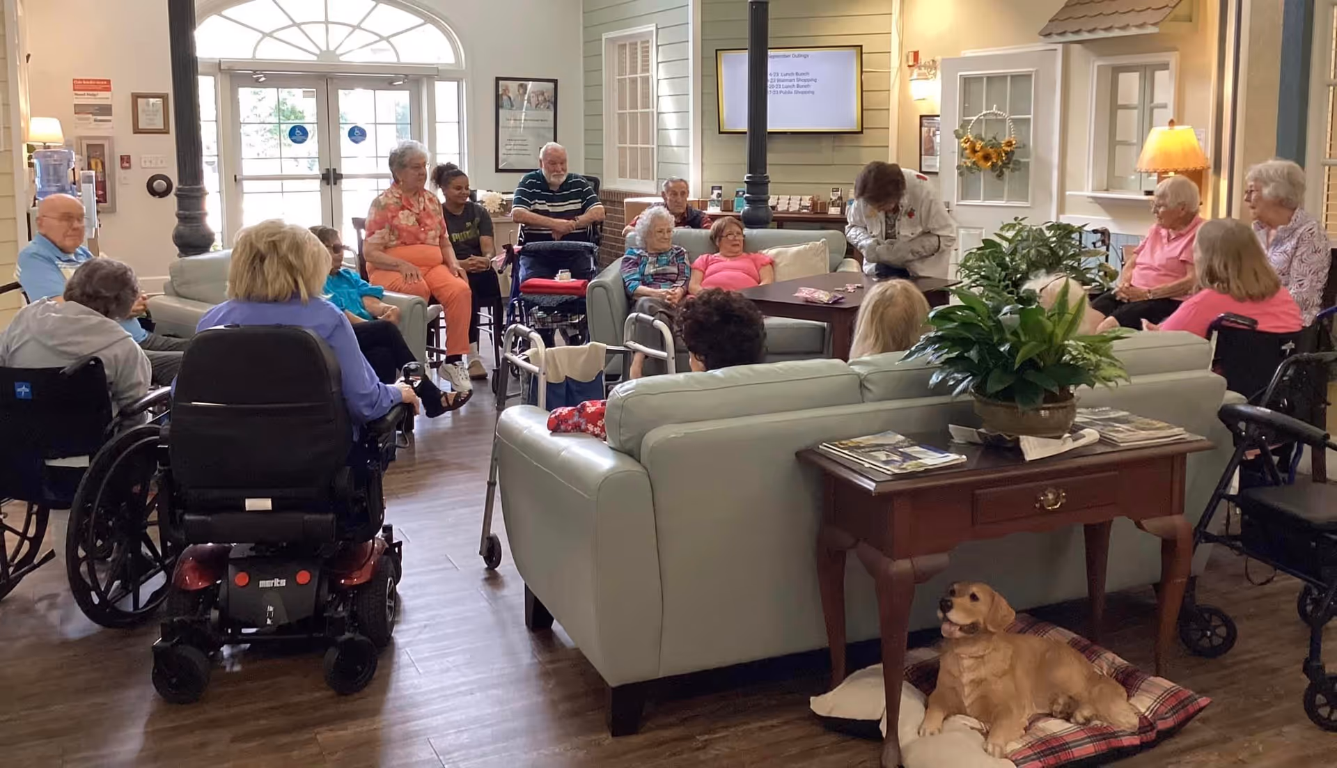 A group of elderly people, some in wheelchairs and some seated on couches and chairs, gathered in a spacious, well-lit common area with wooden floors. A caregiver is interacting with the group. There is a small table with a potted plant and magazines, and a dog lying on a cushion nearby. Large windows and a door with glass panes are visible in the background.