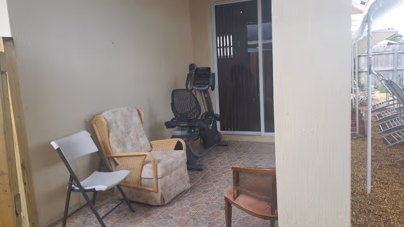 A covered patio area with tiled flooring featuring a white folding chair, a cushioned armchair, a wooden chair, and a stationary exercise bike. The patio opens to an outdoor space with gravel ground and a wooden fence.