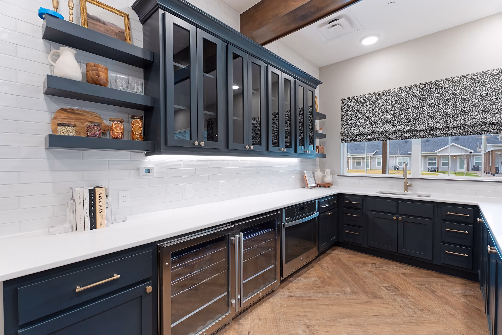 Modern kitchen with dark blue cabinets, white countertops, and a white subway tile backsplash. The kitchen features glass-front upper cabinets, open shelves with decorative items, a built-in oven, and two wine coolers. A large window with a patterned blind overlooks an outdoor area with buildings.