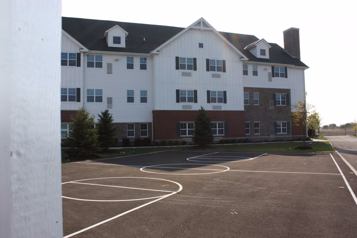Exterior view of a multi-story senior living facility building with white siding, brick and stone accents, multiple windows, and a dark roof. In the foreground, there is an empty outdoor basketball court with white painted lines and a few small trees planted near the building.