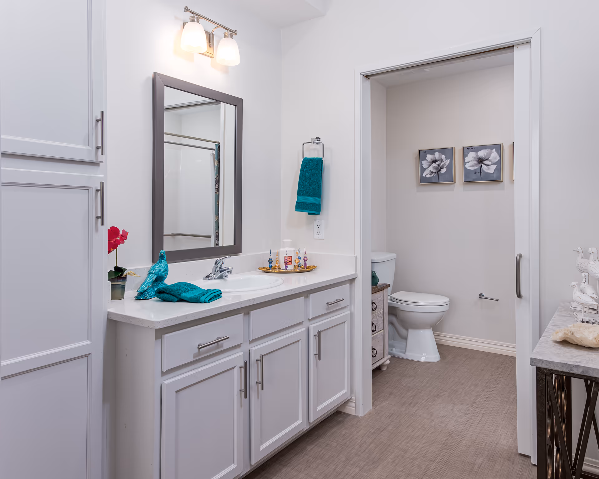 Bright bathroom with a white vanity, mirror and sink, teal towels and decor, and a toilet in a separate alcove.