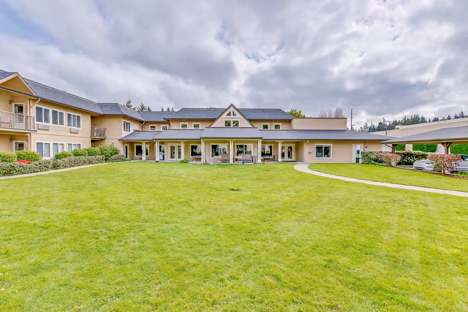 Exterior view of Solstice Senior Living At Normandy Park showing a large green lawn in front of a beige two-story building with multiple windows and a covered porch area under a cloudy sky.