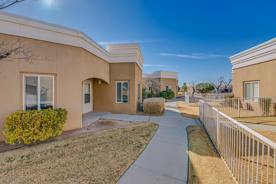 Walkway between single-story beige senior living buildings with shrubs, railings, and a clear blue sky.