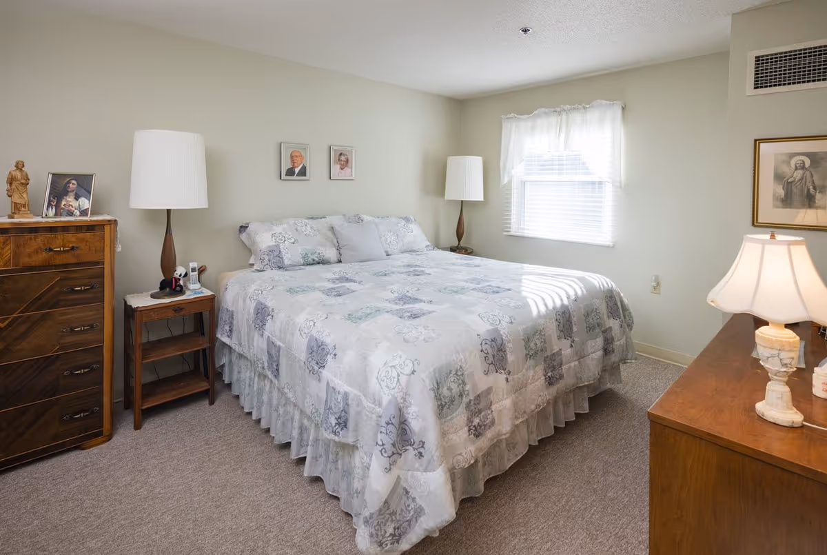A cozy bedroom with a large bed covered in a patterned quilt. There are two bedside tables each with a lamp, a wooden dresser with framed photos and a statue, and a window with sheer white curtains letting in natural light. The walls are light-colored and there are two framed pictures hanging above the bed.