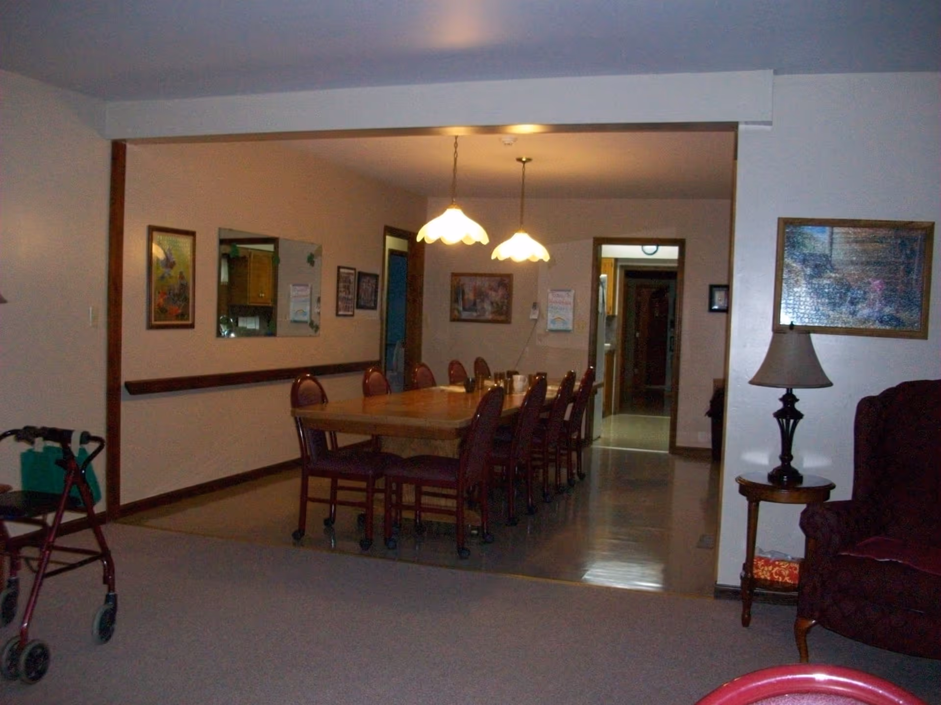 Communal dining room with a long wooden table surrounded by chairs, hanging pendant lights, and a nearby armchair and walker.
