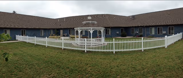 A single-story building with a brown shingled roof and blue exterior walls surrounding a grassy courtyard. In the center of the courtyard is a white gazebo enclosed by a white picket fence. The sky is overcast.