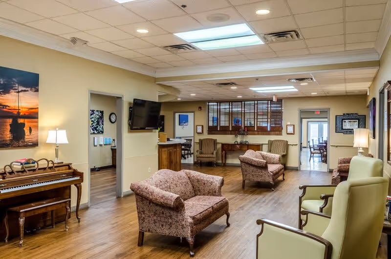 A spacious living room area in a senior living facility with wooden flooring, floral upholstered sofas, green armchairs, a wooden piano with a bench, a wall-mounted TV, and a table with flowers near a window with wooden blinds. The room is warmly lit with lamps and ceiling lights, and there is a doorway leading to another room with a dining area visible in the background.