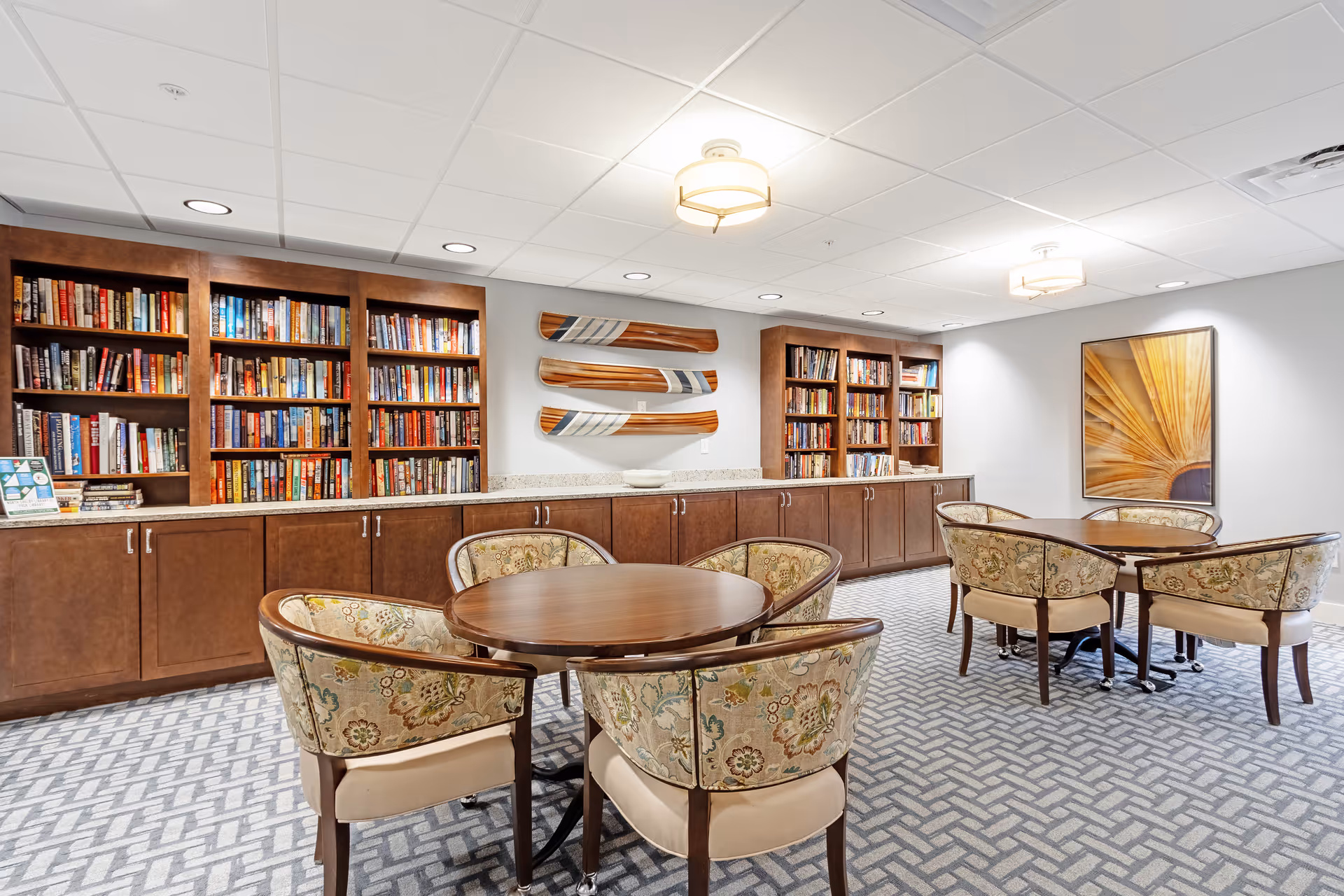 A cozy library or reading room with wooden bookshelves filled with books, two round wooden tables each surrounded by four upholstered chairs with floral patterns, a carpeted floor with a geometric pattern, and three decorative wooden oars mounted on the wall above the cabinets.