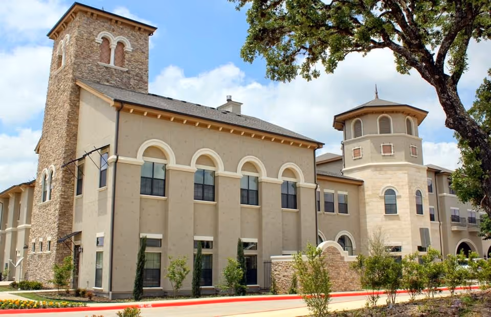 Exterior view of a large senior living facility building with stone and beige stucco walls, arched windows, and two prominent towers. The building is surrounded by small trees and landscaping under a partly cloudy sky.