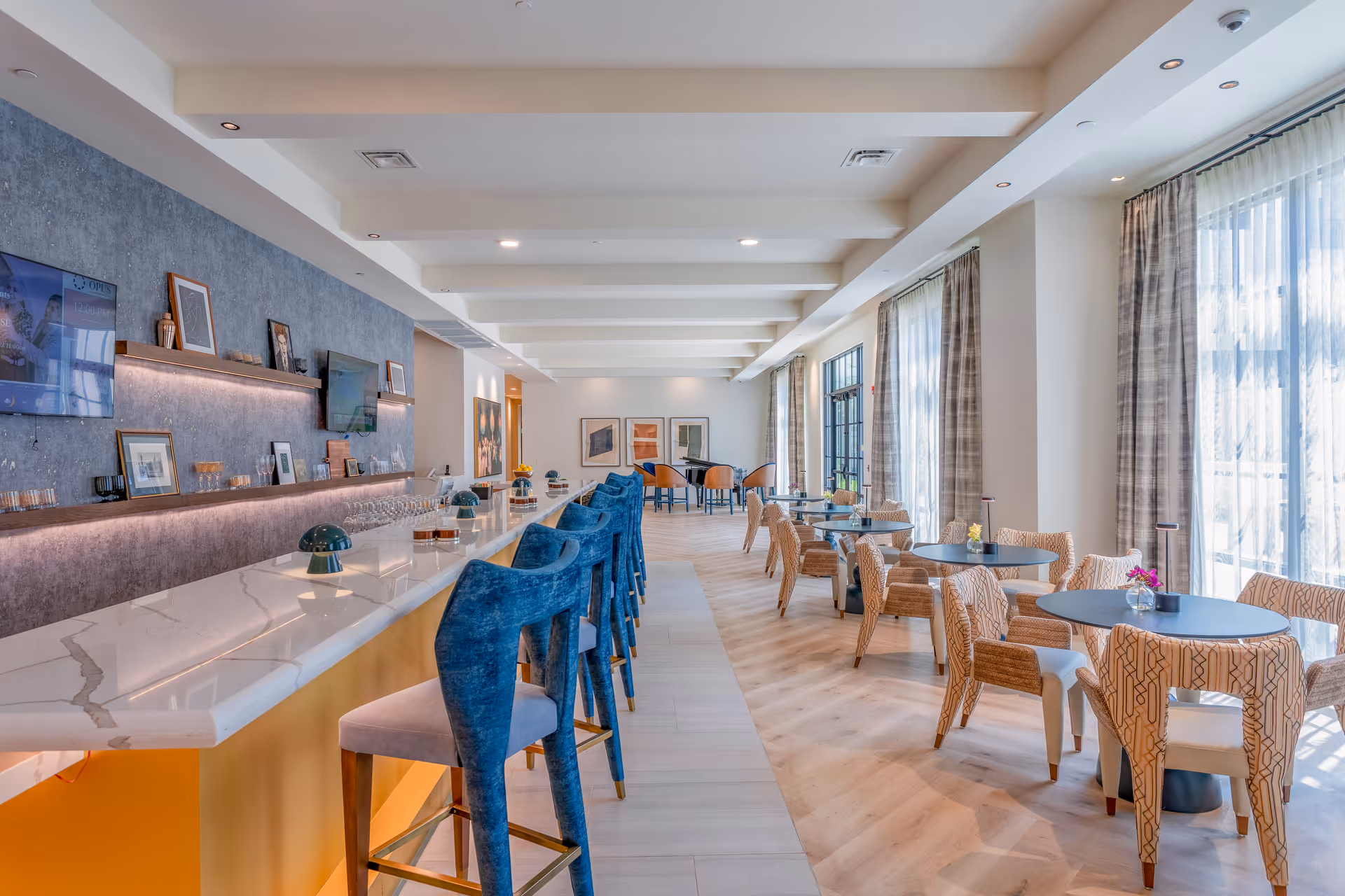 A bright and modern dining area with a long marble countertop bar featuring blue velvet bar stools on the left side. On the right side, there are several round tables with patterned chairs near large windows with sheer curtains allowing natural light to fill the room. The walls have framed artwork and two mounted flat-screen TVs.