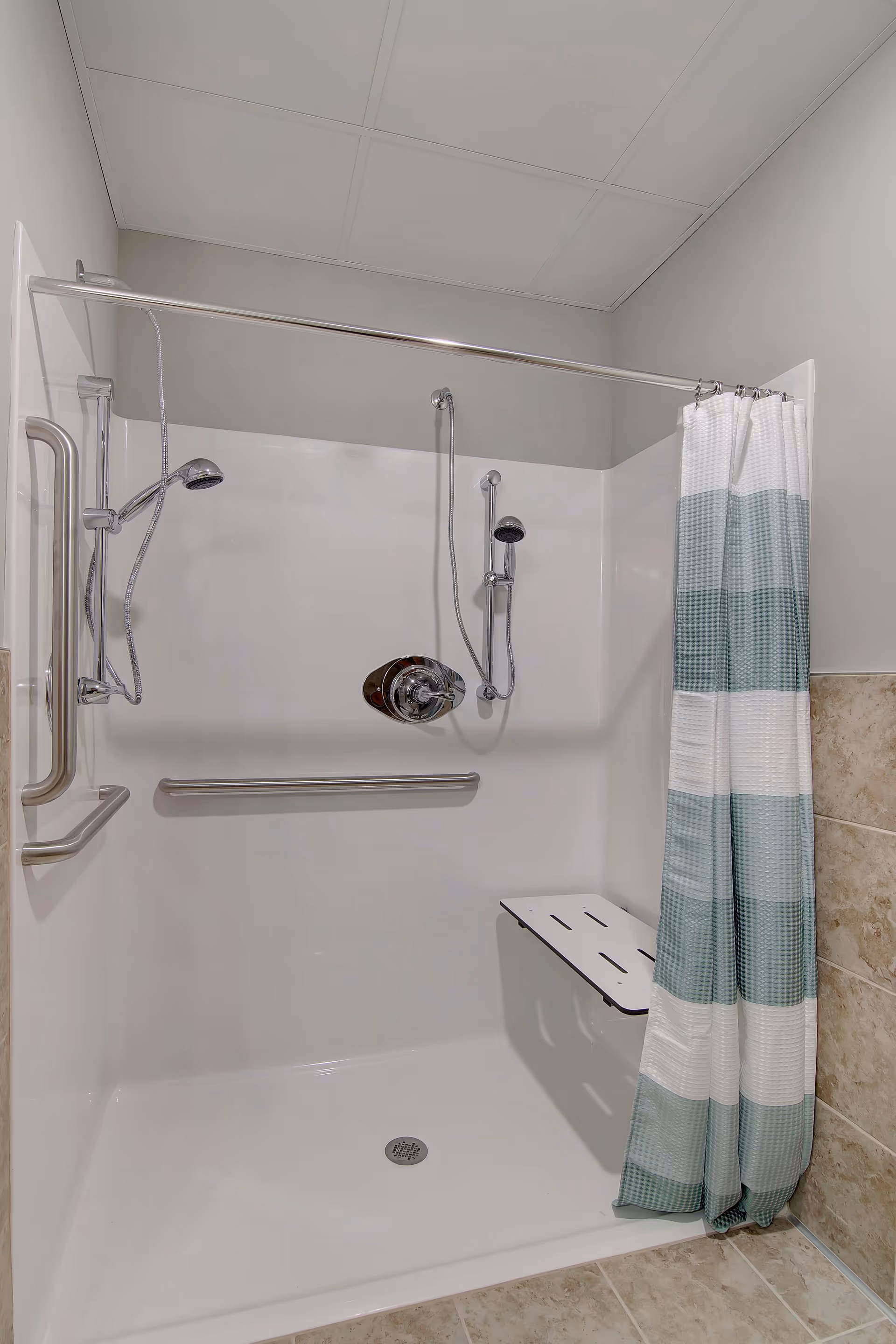 Accessible walk-in shower with grab bars, handheld showerheads, a fold-down seat and a striped shower curtain.