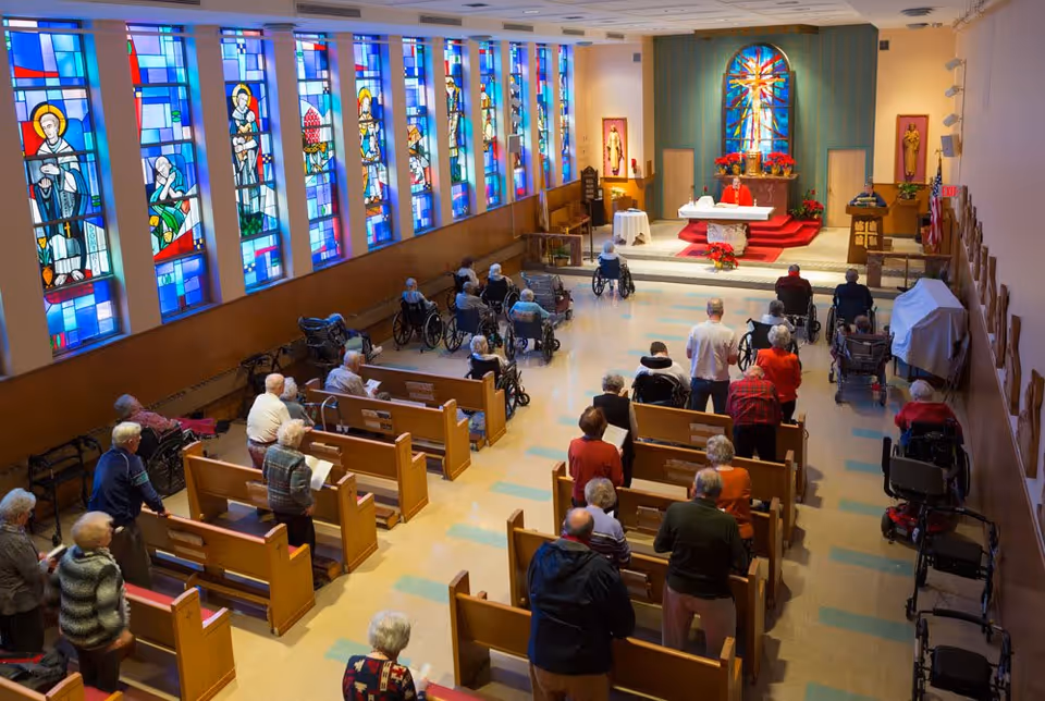 Interior view of a chapel with elderly people attending a service. The chapel has wooden pews, stained glass windows along one wall, and an altar at the front where a person in red robes is standing. Many attendees are seated in wheelchairs, while others stand or sit in the pews.