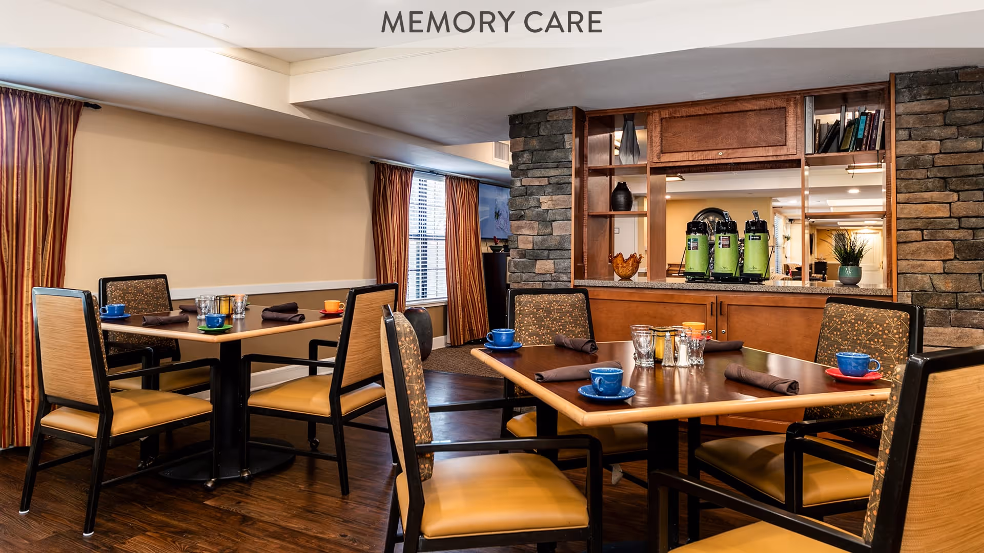 A cozy dining area labeled MEMORY CARE with two square tables set with colorful cups, glasses, and napkins. The room features wooden floors, cushioned chairs, stone accent walls, and a serving window with green beverage dispensers visible in the background. Warm lighting and curtains on the windows add to the inviting atmosphere.