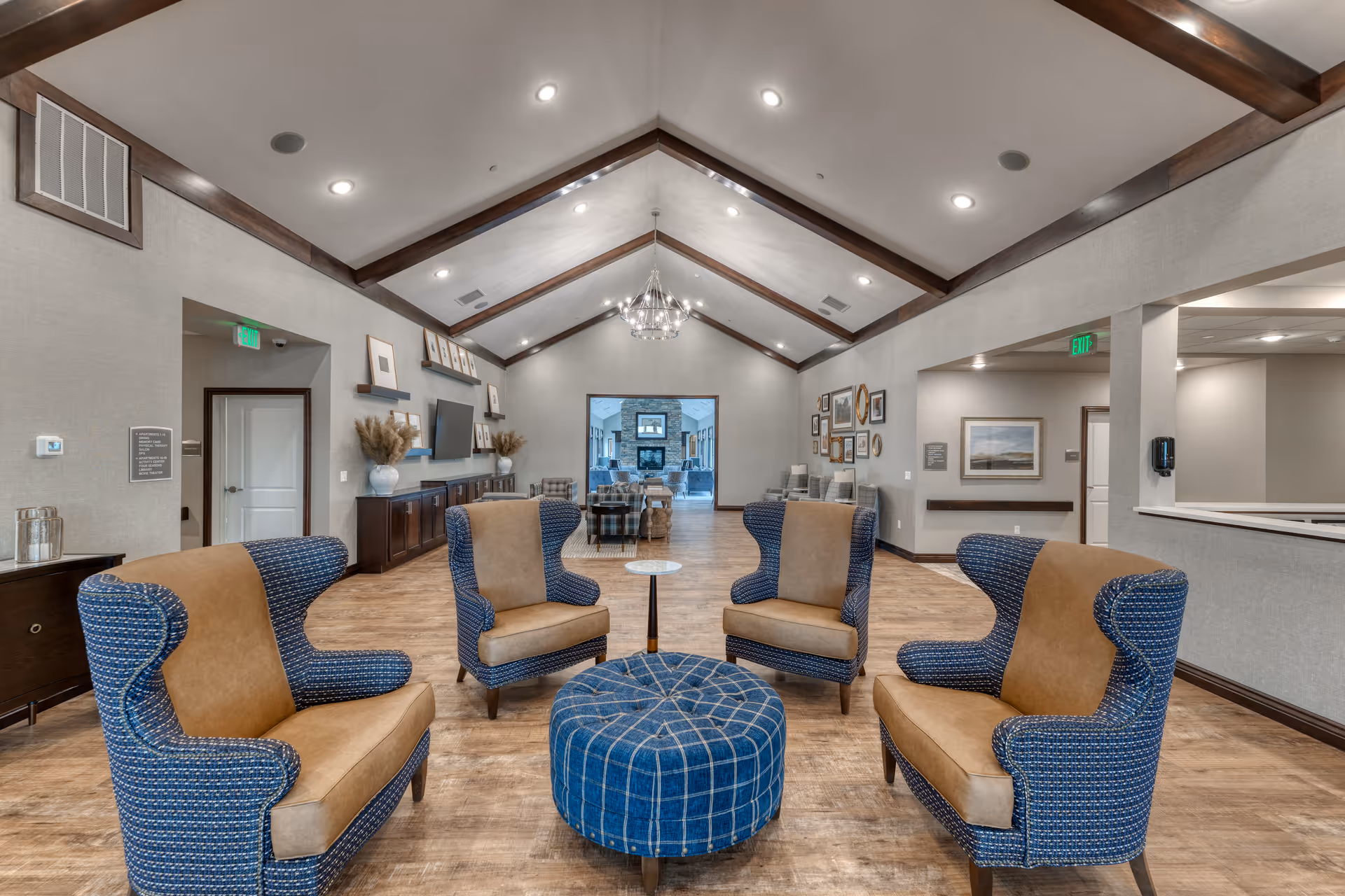 A spacious senior living facility common area with a vaulted ceiling featuring wooden beams and a chandelier. The room has four blue and tan upholstered armchairs arranged around a round blue ottoman on a wooden floor. The walls are decorated with framed pictures and a flat-screen TV mounted above a dark wooden cabinet. There are doorways and open spaces leading to other rooms in the background.