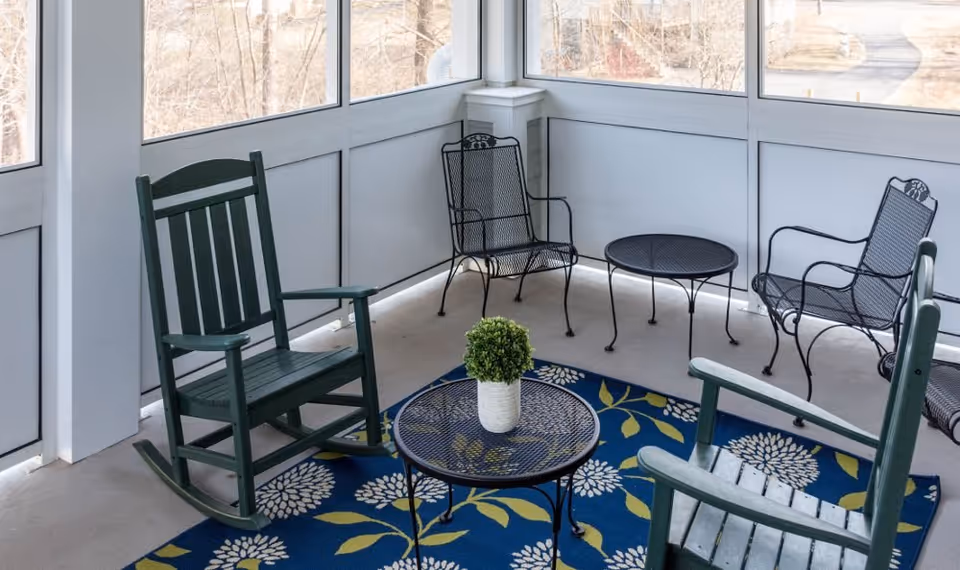 A screened-in porch area with two green wooden rocking chairs, two black metal chairs, and two black metal tables. A blue rug with white and yellow floral patterns is on the floor, and a small potted plant is on one of the tables. The porch overlooks a natural outdoor setting with trees and a pathway.