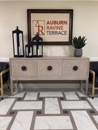 A decorative console table with three drawers featuring circular handles, flanked by two chairs with wooden arms and black cushions. On top of the table are three black lanterns with white candles inside and a gray pot with a green plant. Above the table is a framed sign that reads 'Auburn Ravine Terrace'. The floor has a patterned tile design in white and brown tones.