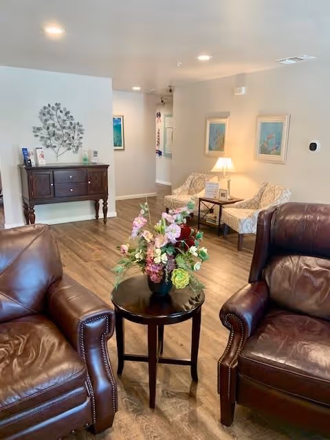 A cozy interior lounge area with two brown leather armchairs facing a small round table with a floral arrangement. In the background, there is a wooden sideboard with decorative items and two patterned armchairs next to a small table with a lamp. The walls are light-colored with framed artwork and the floor is wood.