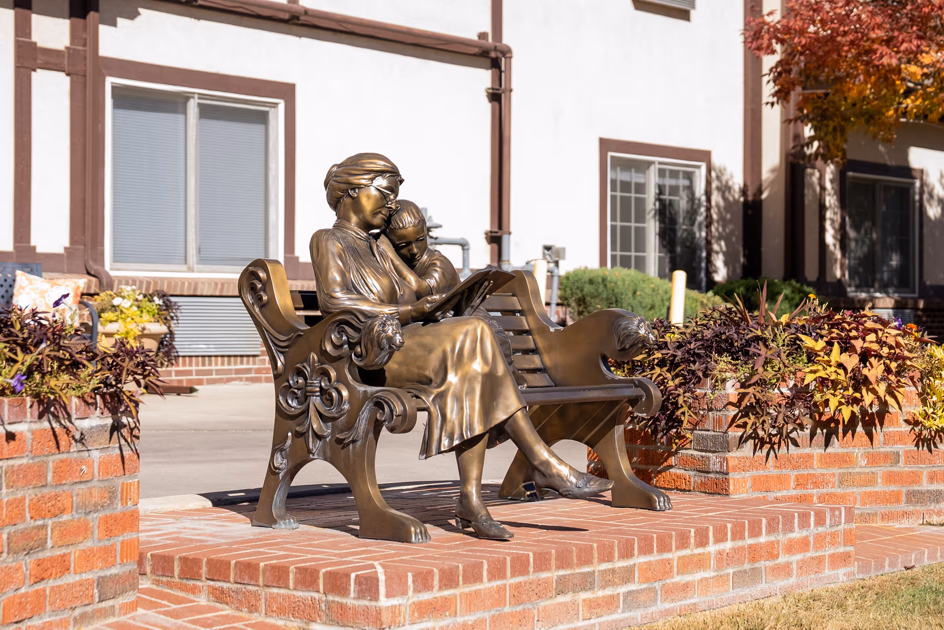 Bronze statue of a woman reading a book to a child while sitting on an ornate bench, placed on a brick platform with flower beds on either side, in front of a building with windows and brown trim.