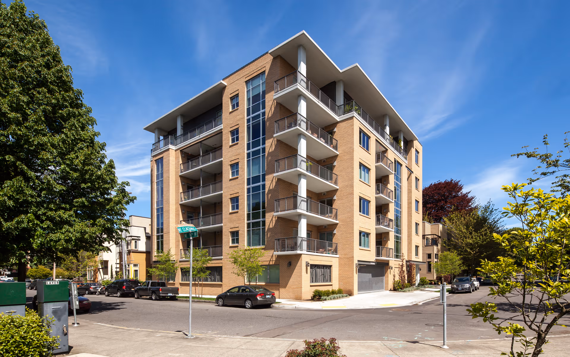 Six-story modern brick apartment building with balconies on a sunny, tree-lined street corner.