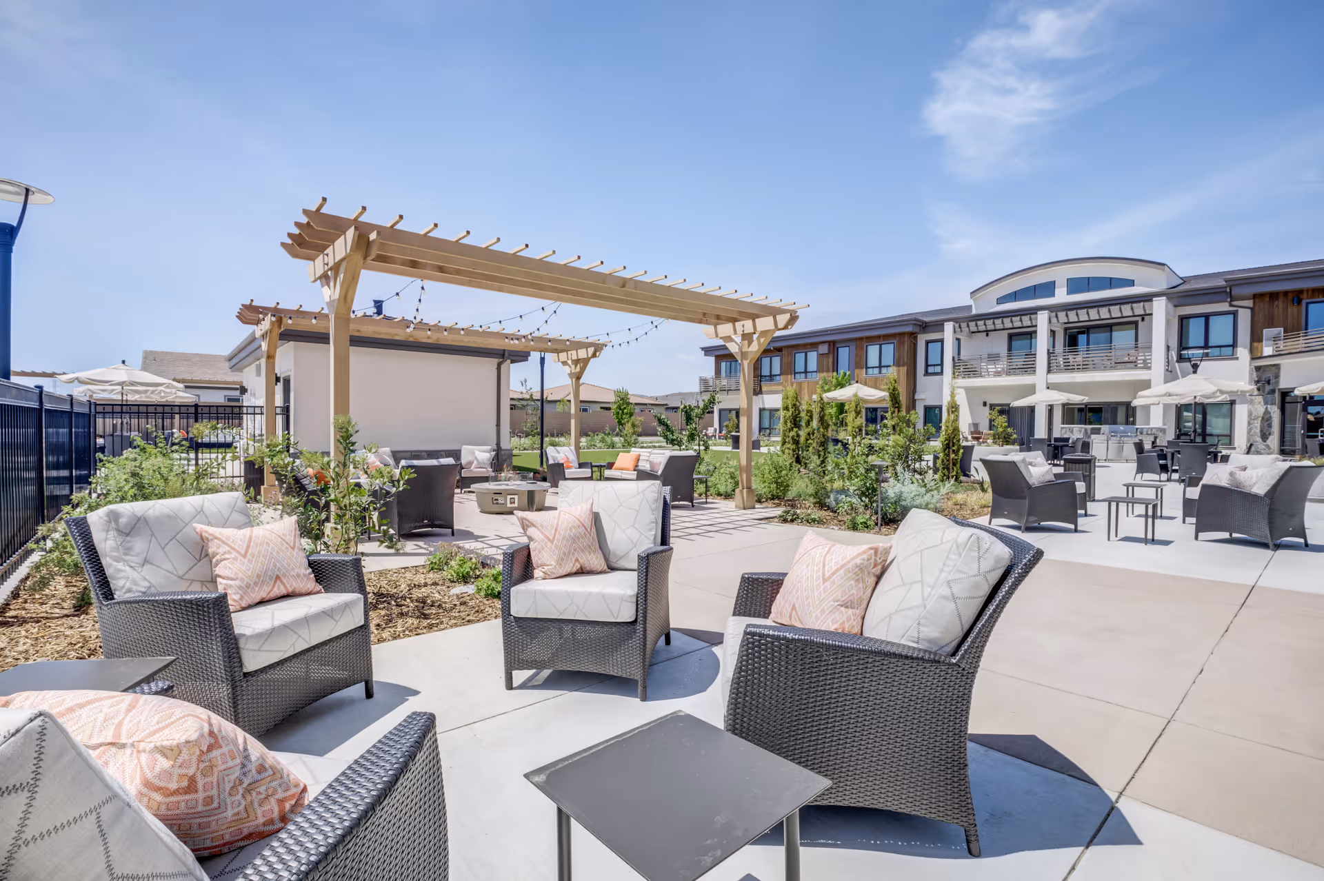 Outdoor courtyard with wicker lounge chairs, cushions, a pergola with string lights, and the apartment building in the background.
