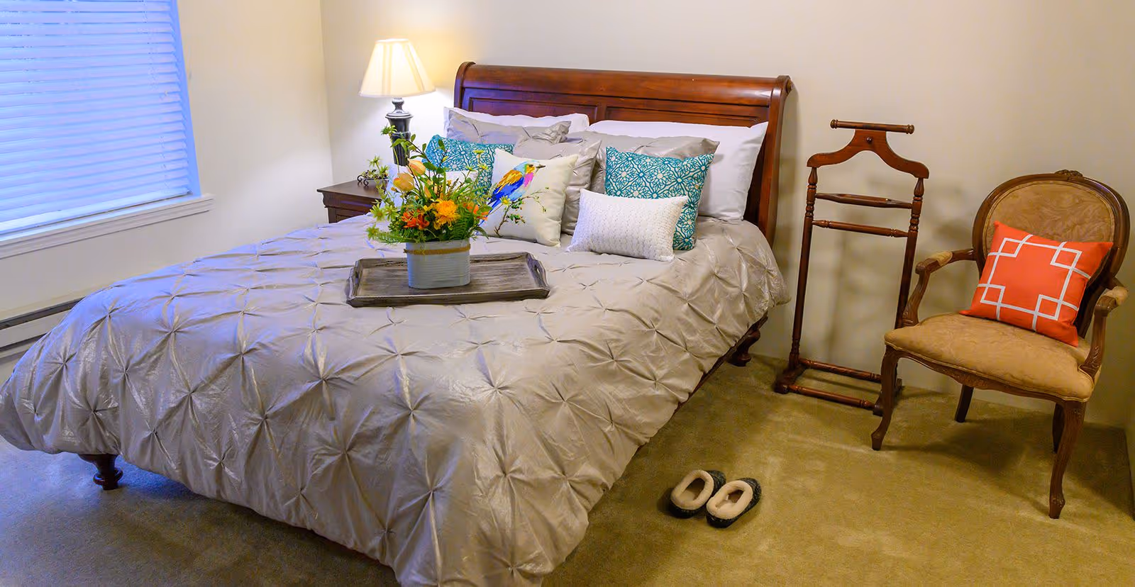 A neatly made bedroom with a wooden sleigh bed, decorative pillows and a tray of flowers, plus a chair and slippers on the carpet.