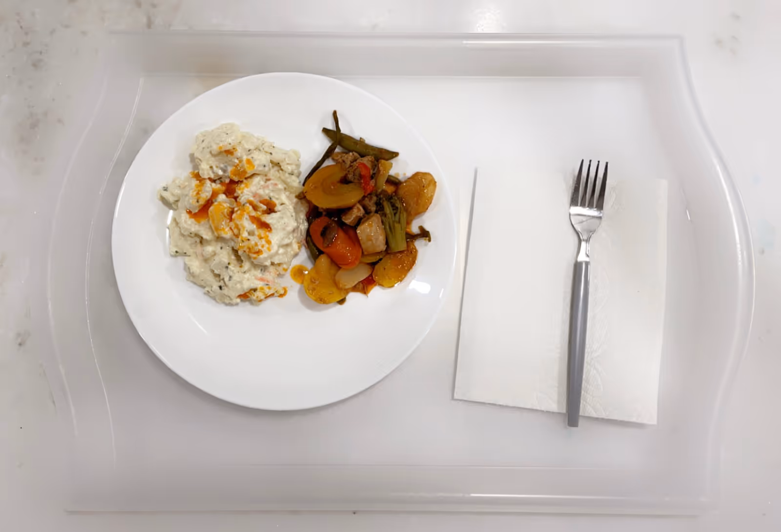 A white plate on a clear tray containing a serving of creamy mashed potatoes with gravy and a portion of cooked mixed vegetables and meat. Next to the plate is a white napkin with a fork placed on top.