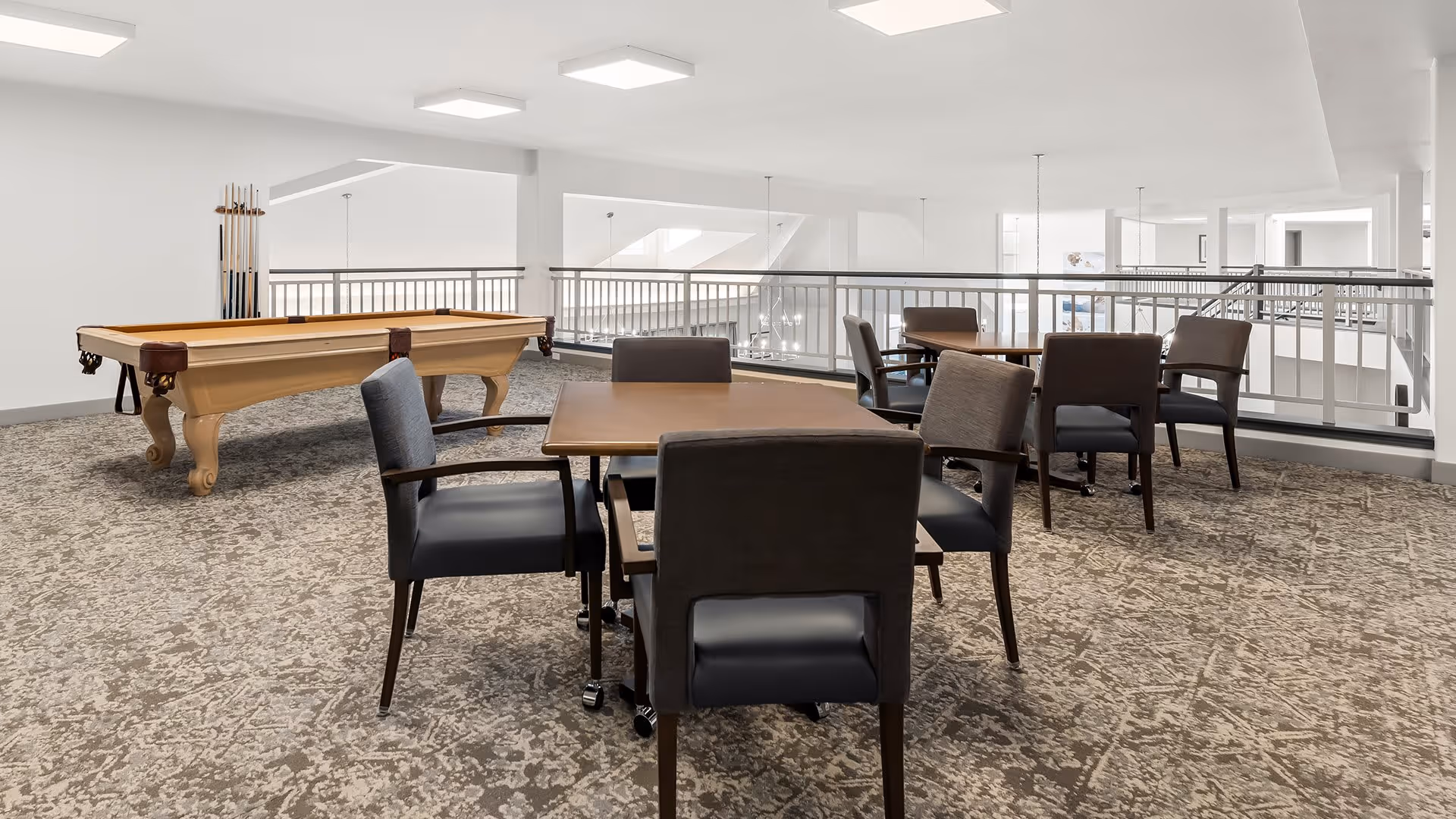 A bright, spacious common area with carpeted floors featuring several tables and chairs arranged for seating. A pool table with cues is positioned near the back left side. The area is surrounded by white walls and railings overlooking a lower level, with ceiling lights providing illumination.