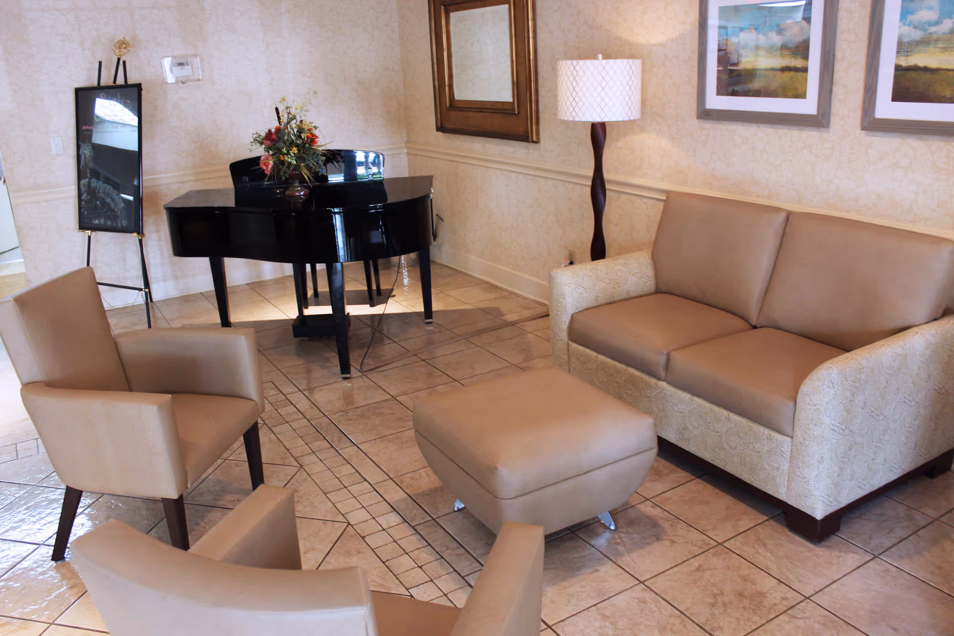 A cozy sitting area with a beige loveseat, two beige armchairs, and a matching ottoman on a tiled floor. A black grand piano with a floral arrangement on top is in the corner, next to a floor lamp and framed artwork on the walls.