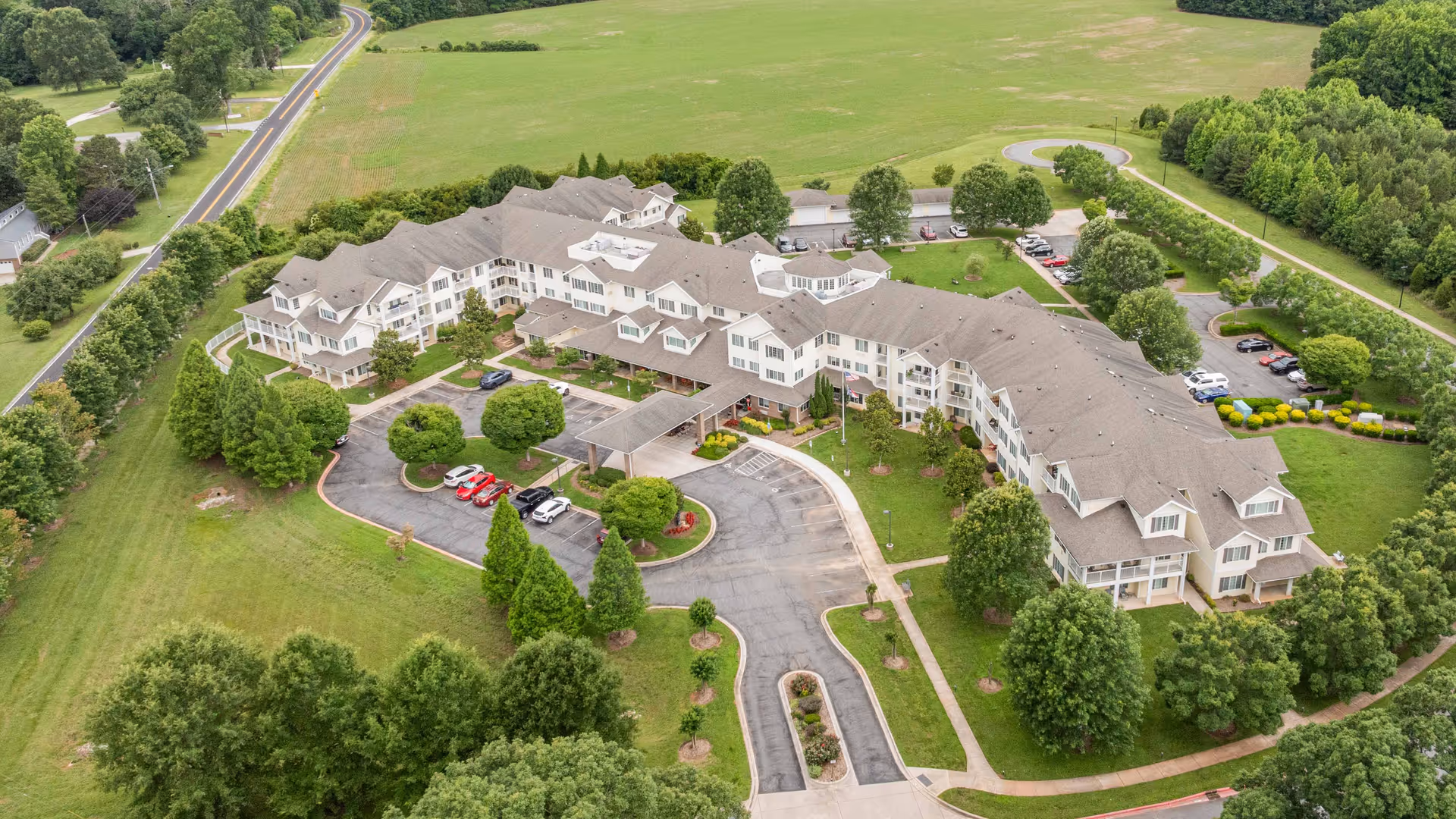 Aerial view of Oak Park Retirement, a large multi-wing building surrounded by green lawns, trees, and parking areas. The building has multiple floors with a gray roof and white exterior walls. There is a driveway leading to a covered entrance and several cars parked in the parking lot. The surrounding area includes open fields and a road.