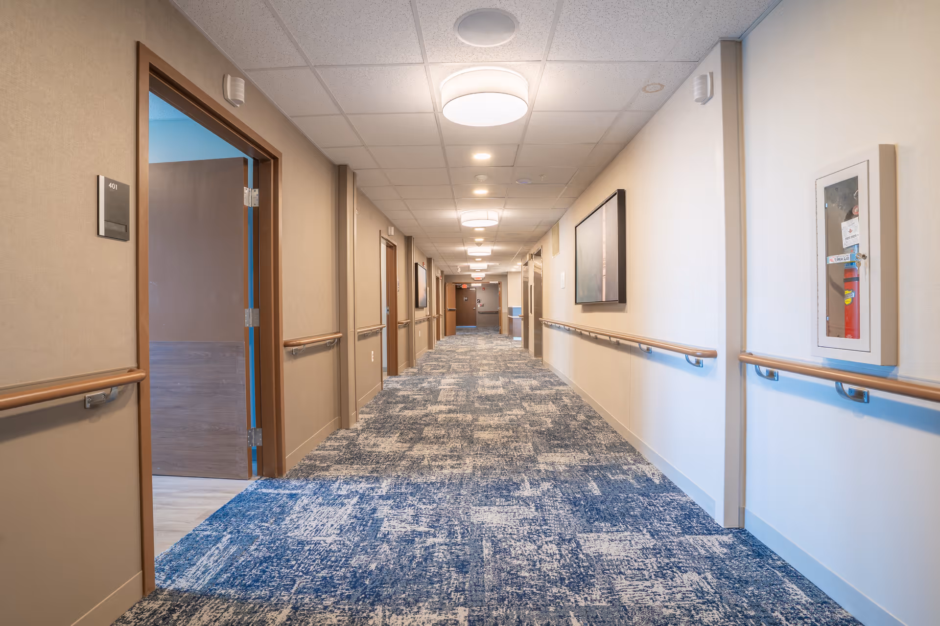 A long, well-lit hallway in a senior living facility with blue patterned carpet, beige walls, handrails on both sides, several closed doors, and ceiling lights. A fire extinguisher is mounted on the right wall inside a glass case.