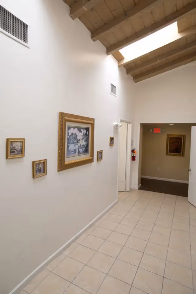 A bright interior hallway with a tiled floor, white walls decorated with framed pictures, and a wooden beamed ceiling with skylights leading to an open doorway.