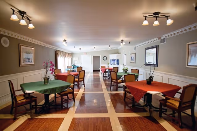 A dining room in a senior living facility with several tables covered in red and green tablecloths, each surrounded by wooden chairs with cushions. The room has wood-patterned flooring with a light border, beige walls with white wainscoting, framed artwork, and ceiling lights. There are windows with curtains on the left side and a clock on the far wall.